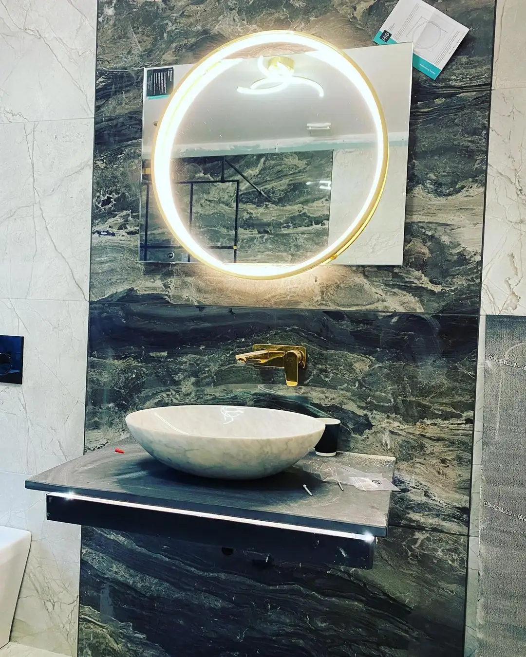 Modern bathroom with round vessel sink, gold wall-mounted faucet, and LED-lit circular mirror on dark marble wall