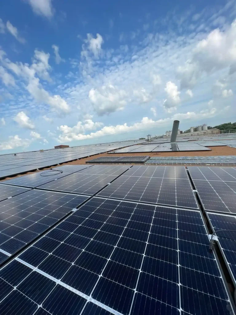 Large rooftop solar panel array under partly cloudy sky, with industrial buildings and cylindrical tanks in background