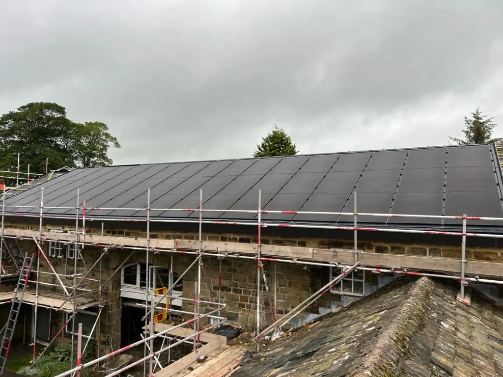 Sloped rooftop fully covered with grid-pattern solar panels under cloudy sky, with trees and construction equipment in background