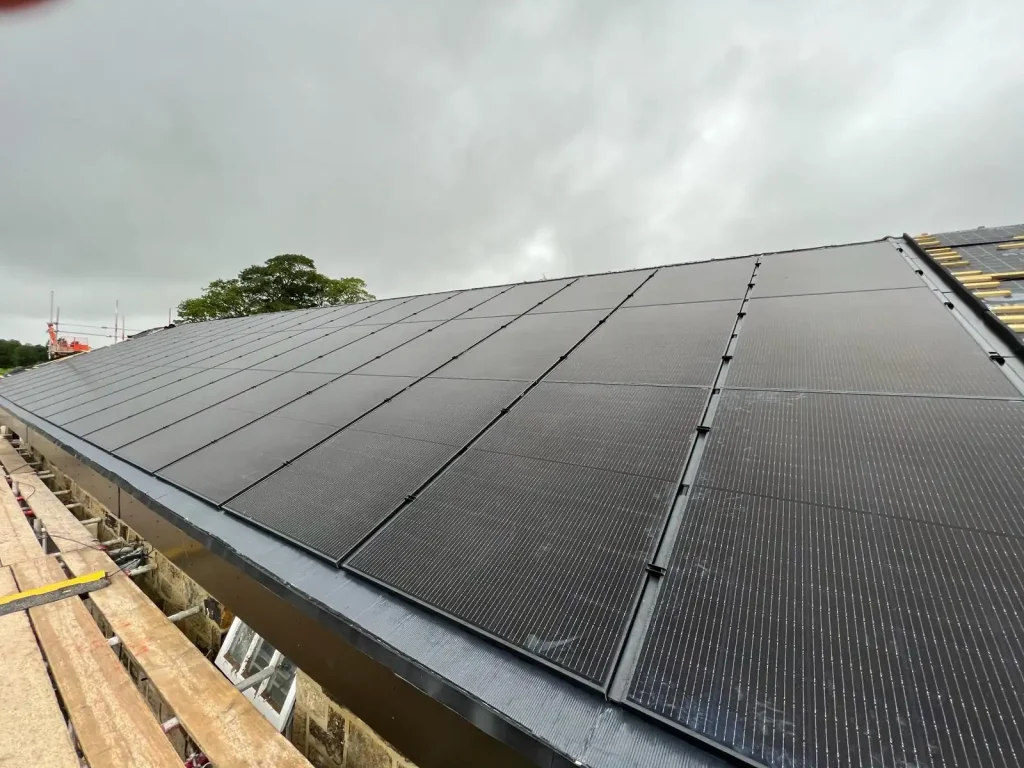Sloped rooftop fully covered with grid-pattern solar panels under cloudy sky, with trees and construction equipment in background